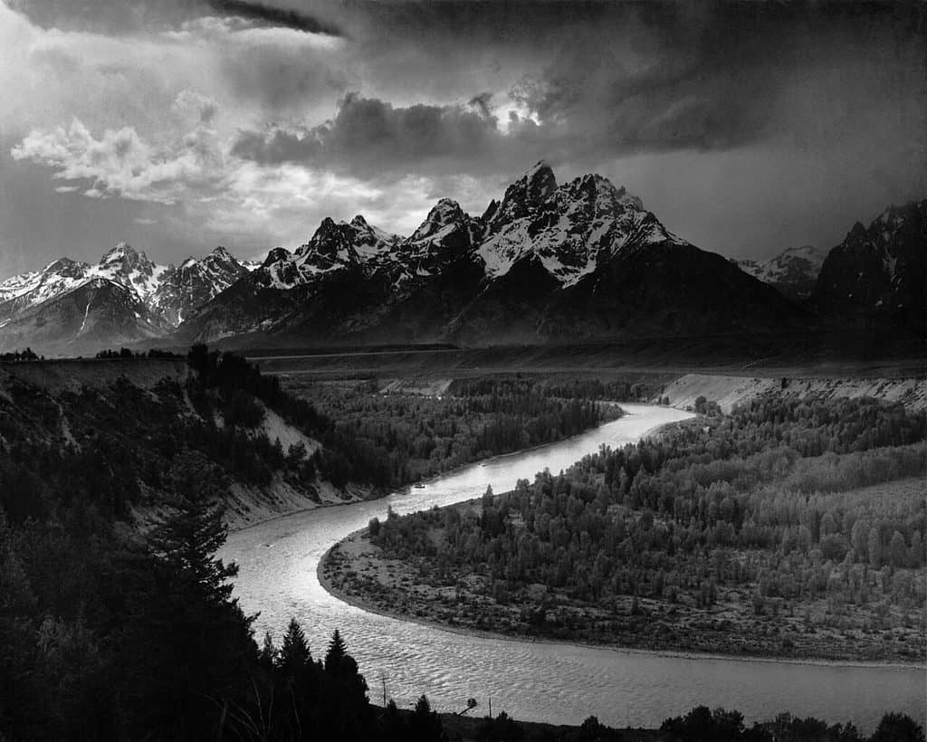Svartvitt landskap där Snake River slingrar sig som ett ljust S genom en mörk barrskogsdal, med snöklädda bergstoppar och ett dramatiskt molnband bakom.