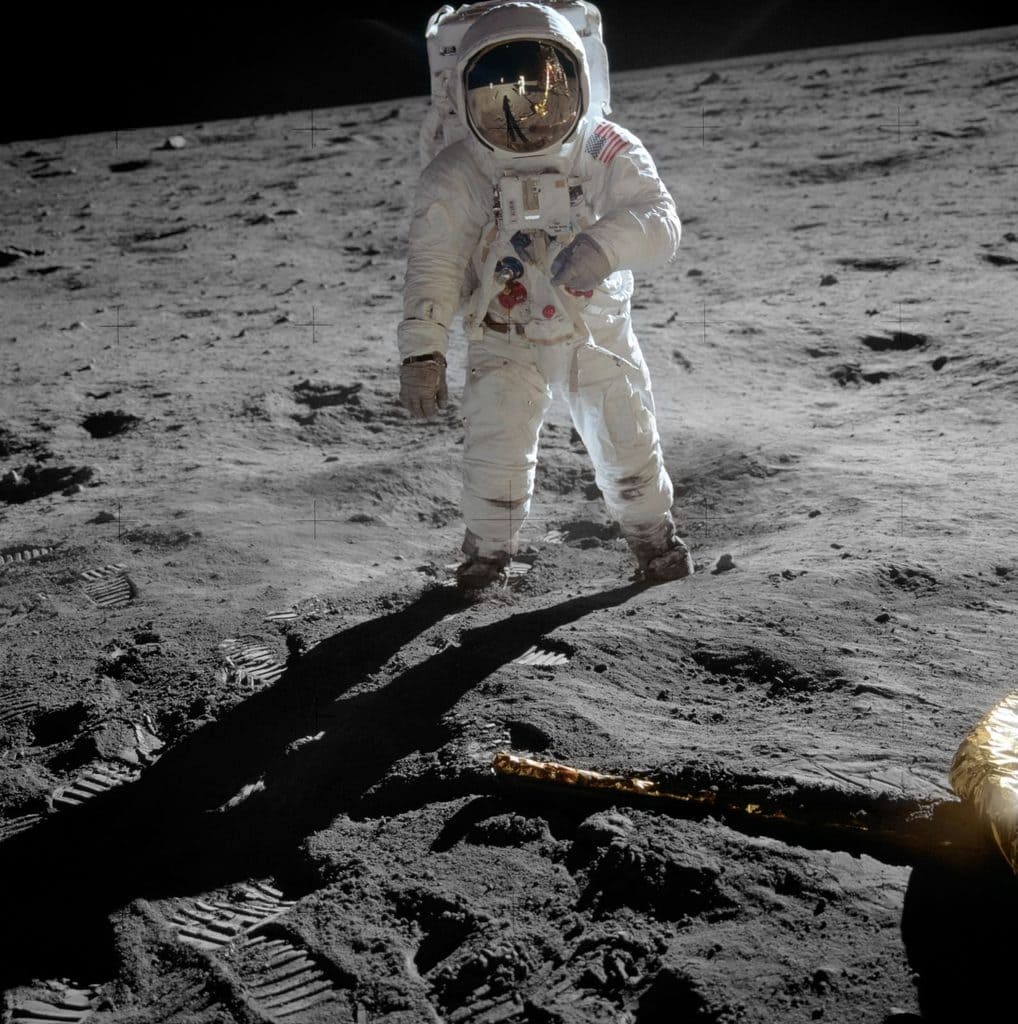 En astronaut i vit rymddräkt står vänd mot kameran på månytan, något lutad, med armarna längs sidorna. I det guldade visiret speglas fotografen, månlandaren och det upplysta landskapet. Överkanten av ryggsäcken är avskuren av bildkanten, och bakom hjälmen finns bara en smal remsa svart himmel.
