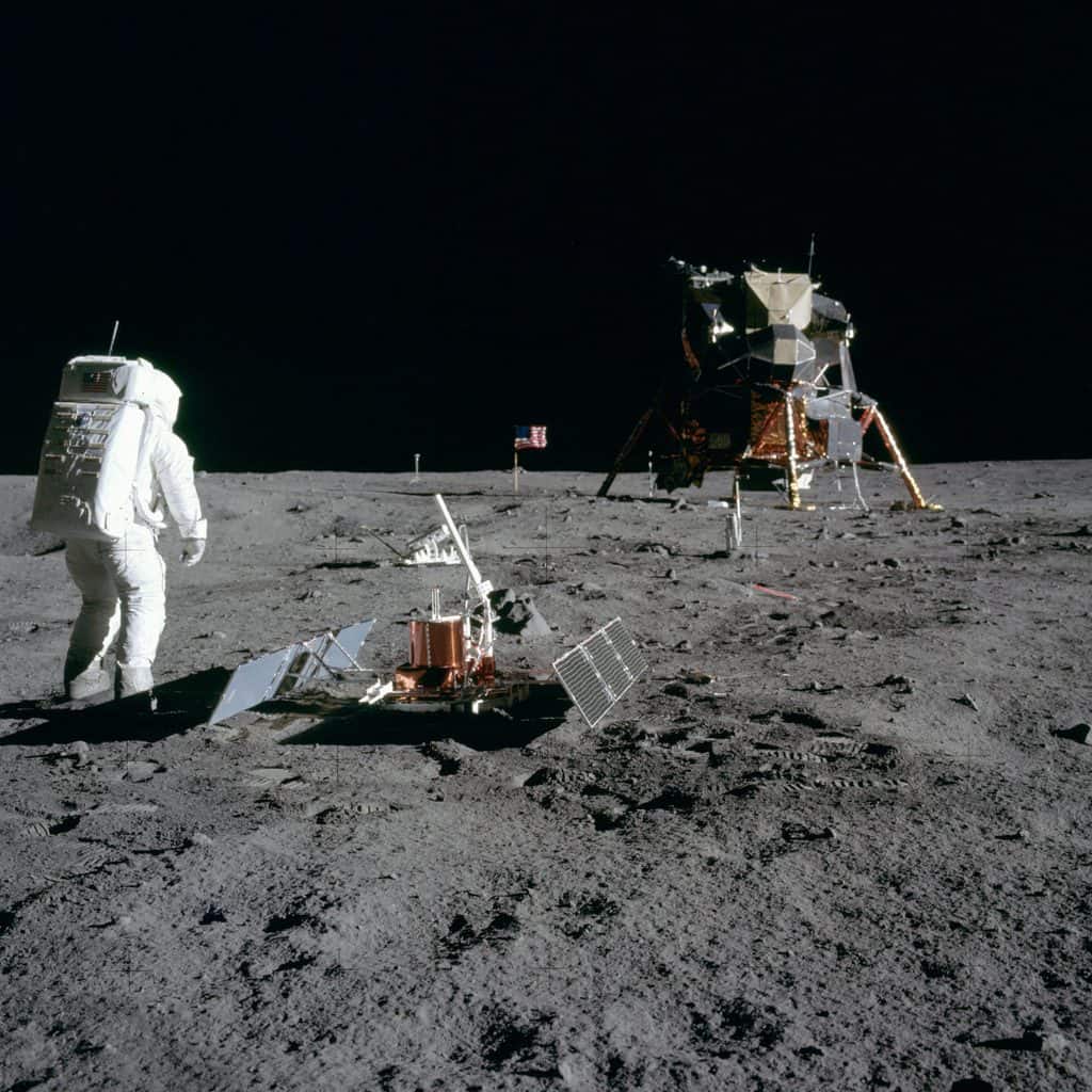 Astronaut i vit rymddräkt går åt vänster över månytan. I förgrunden står ett kopparfärgat instrument med två utfällda solpaneler, snett bakom det ligger en tiltad reflektorpanel, längre bort står en amerikansk flagga, och till höger står månlandaren Eagle på fyra ben under svart himmel.