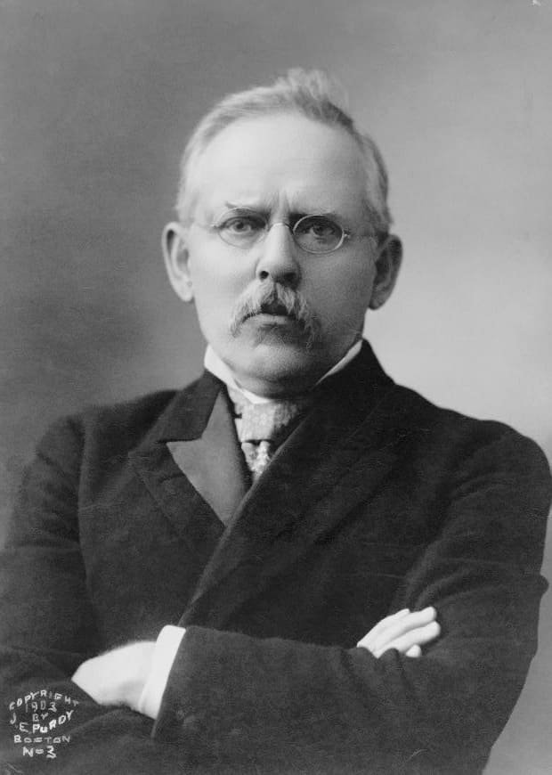 Porträtt av Jacob Riis i mörk kavaj med armarna i kors, glasögon, mustasch och kort ljust hår, halvfigur mot grå bakgrund