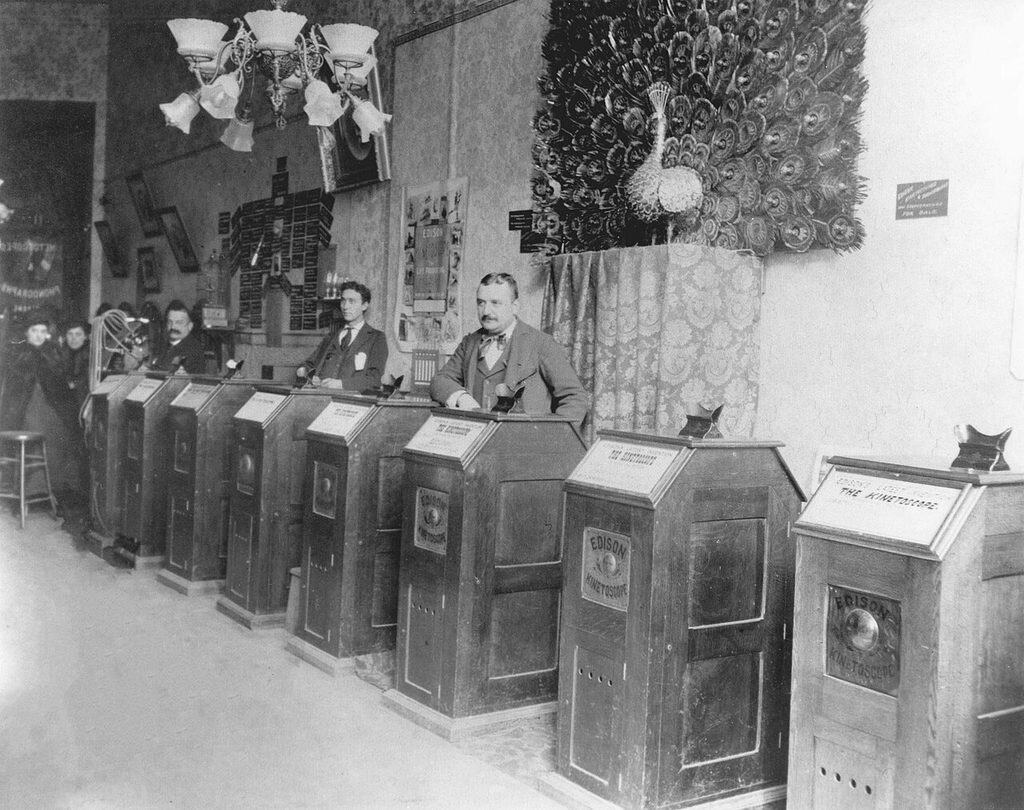 Interiör med en rad höga träskåp som löper in i bilddjupet. Varje skåp har texten EDISON KINETOSCOPE, ett mässingsbeklätt titthål på locket och ett kort på framsidan. Tre män i kostym står bakom raden, en gaskrona hänger i taket och en uppstoppad påfågel med spretande stjärt sitter mot bakväggen.