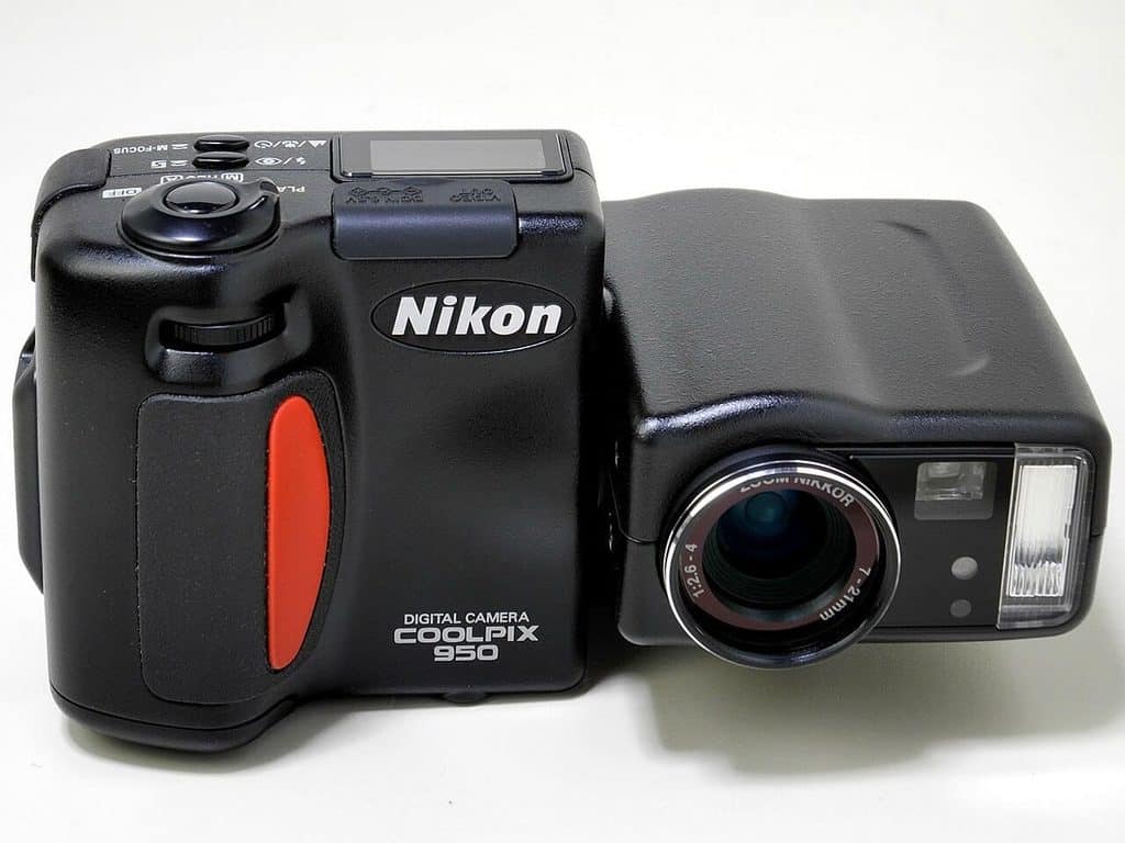 Svart digitalkamera i två delar. Den främre delen har handgrepp, ratt, en liten display på ovansidan och texten Nikon samt DIGITAL CAMERA COOLPIX 950, med ett rött gummifält på framsidan. Den bakre delen är vriden i vinkel mot den främre och bär objektivet med texten ZOOM NIKKOR.