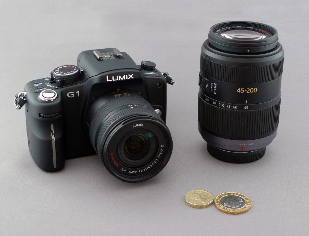 Panasonic Lumix G1 med normalzoom, bredvid ett längre telezoomobjektiv och några mynt som visar kamerans ringa storlek.