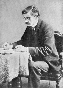 Man med glasögon och mustasch sitter i en karmstol vid ett litet dukat bord och skriver med penna på ett papper, svartvitt studiofotografi från 1880-talet.