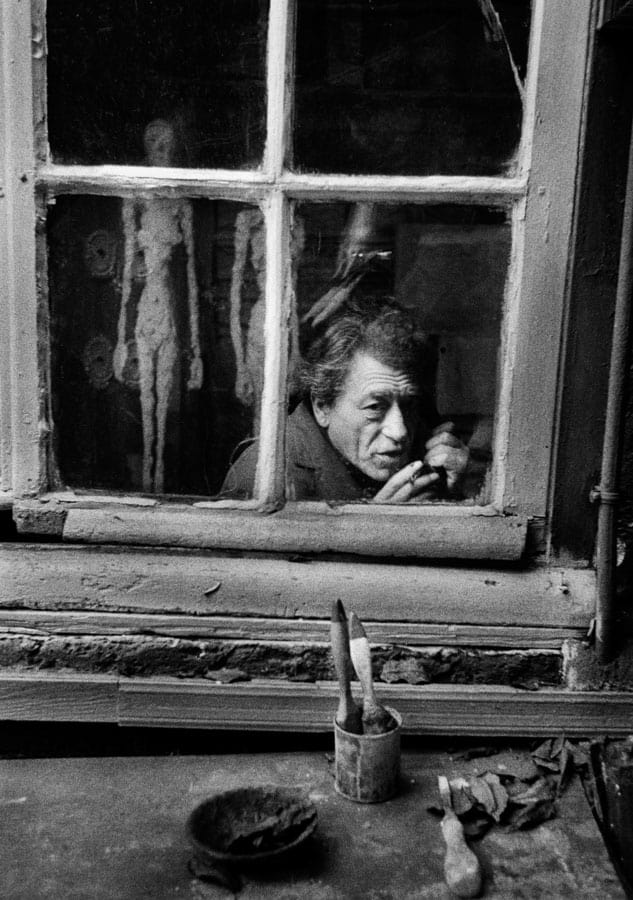 Konstnären Alberto Giacometti bakom en smutsig spröjsad fönsterruta, med spröda figurer skymtande i glaset och penslar på fönsterbrädan.