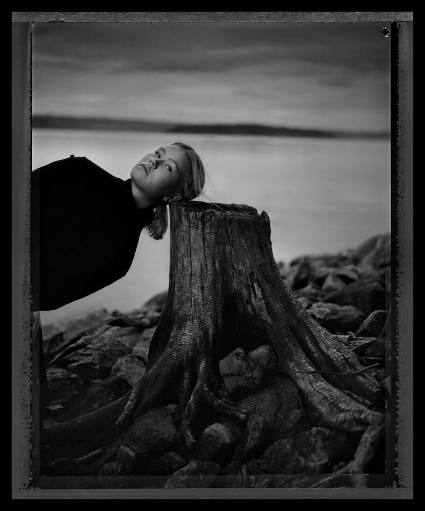 Ung flicka i mörk tröja lutar bakhuvudet mot en stor stubbe på en stenig strand med vatten och låg horisont bakom