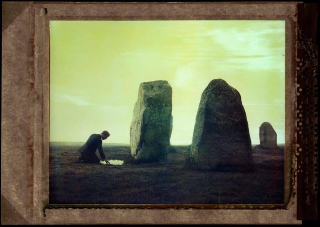 Knäböjande gestalt vid resta stenar i skymning, polaroid med blekt gulgrön ton
