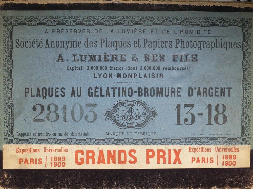 Blå tryckt etikett på ett kartonglock, inramad av ett ornament. Fransk text: Société Anonyme des Plaques et Papiers Photographiques, A. Lumière & ses fils, Lyon-Monplaisir, Plaques au gélatino-bromure d'argent. I mitten ett ovalt varumärke, flankerat av stora siffror. Nedtill en ljus remsa med röd text om Grands Prix vid världsutställningarna i Paris 1889 och 1900.