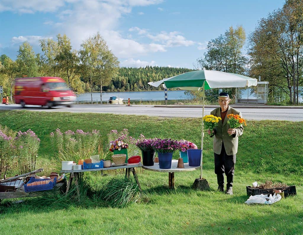 Äldre man i keps håller upp två blombuketter vid ett vägkantsstånd med parasoll, en röd skåpbil far förbi