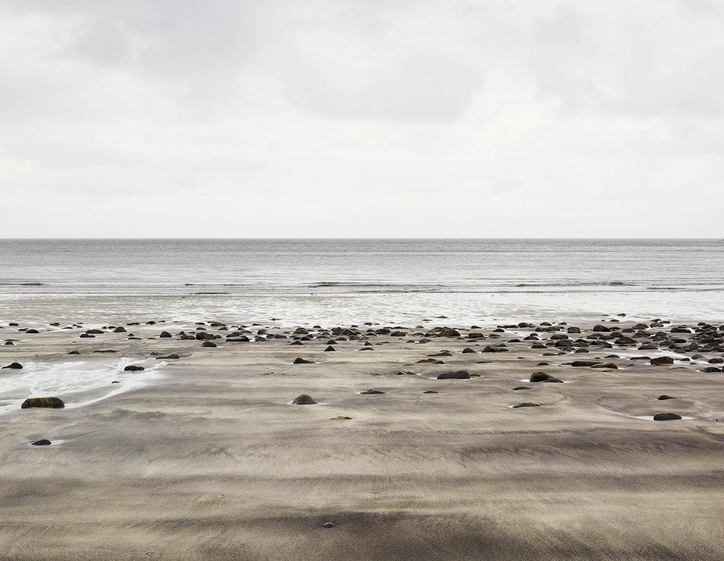 Spridda stenar på en bred sandstrand vid lågvatten, platt hav och grå himmel vid Talisker Bay