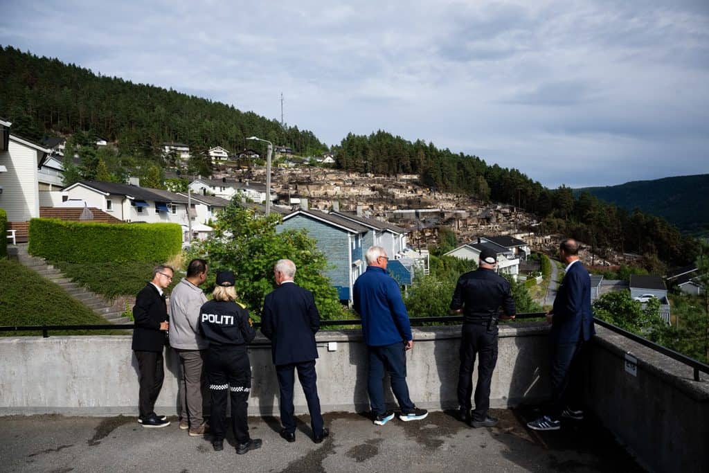 Kronprins Haakon och sex andra personer står vid ett räcke och ser ut över nedbrunna hus i Krokstadelva.