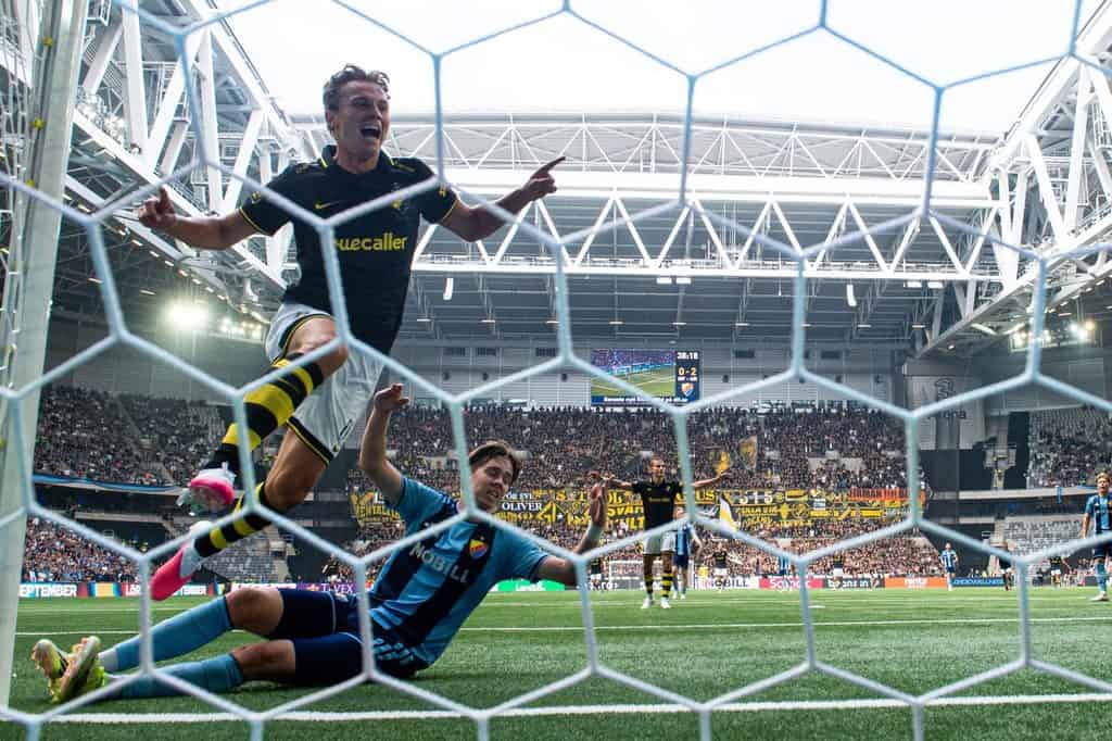 Johan Hove jublar med utsträckta armar, sedd genom målnätet, medan en Djurgårdsspelare ligger på gräset.