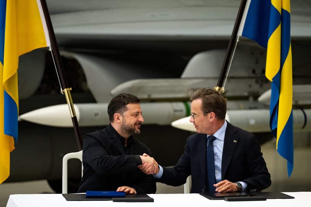 Volodymyr Zelenskyj och Ulf Kristersson skakar hand vid ett bord mellan ukrainska och svenska flaggor, med ett Gripenplan bakom.