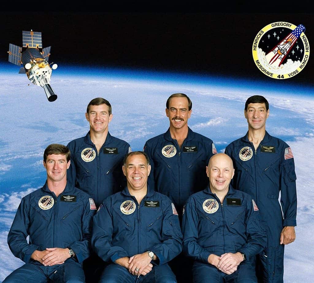 ex astronauter i mörkblå flygoveraller, tre stående bakom tre sittande, mot en bakgrund av jordens molntäcke och en satellit med utfällda solpaneler. Uppe till höger uppdragsmärket för STS-44