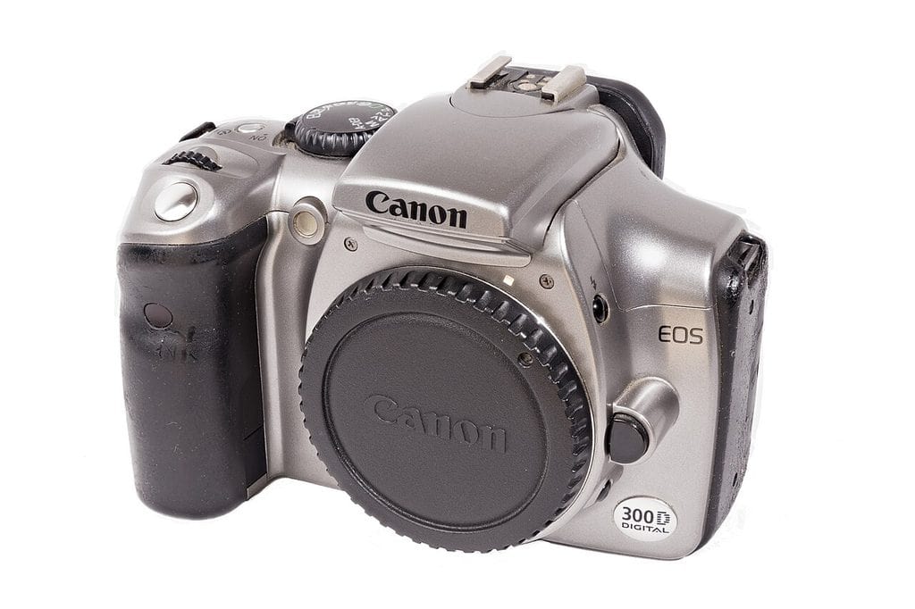 Canon EOS 300D snett framifrån mot vit botten, hus utan objektiv och med svart bajonettlock. Silverfärgat plasthölje med svart gummigrepp, lägesratt uppe till vänster, blixtsko på toppen och en oval etikett märkt 300D DIGITAL nere till höger
