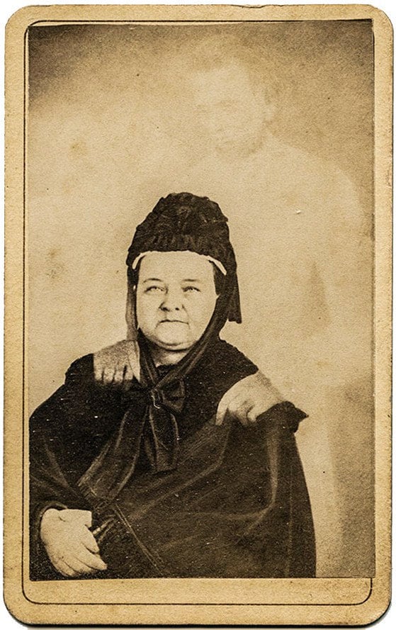 Mary Todd Lincoln sittande i svart sorgklänning, med en blek genomskinlig gestalt av Abraham Lincoln bakom sig som vilar händerna på hennes axlar.