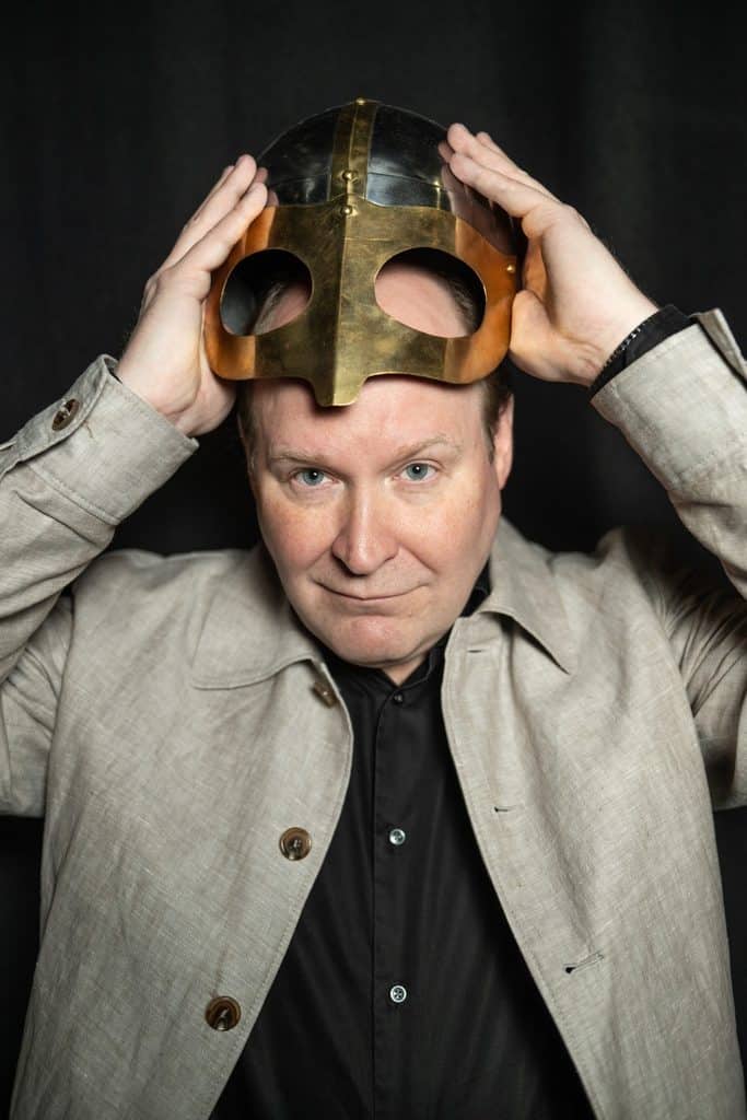 Henrik Dorsin håller en vikingahjälm ovanför pannan, mot svart bakgrund