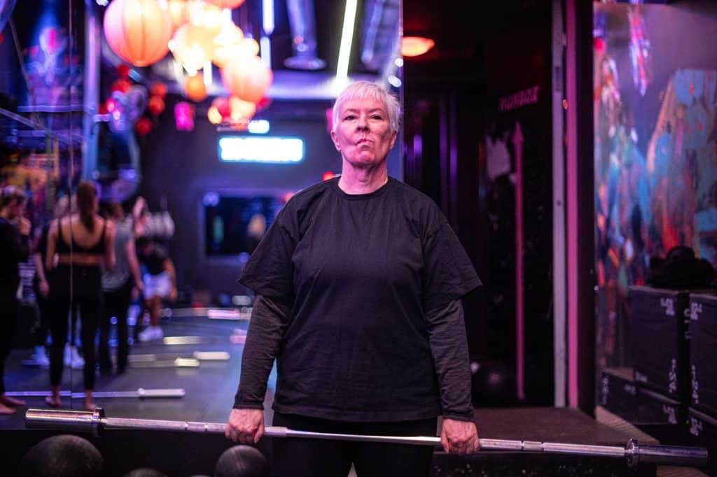 Mona Sahlin står med en skivstång framför sig i ett gym med papperslyktor och neonljus