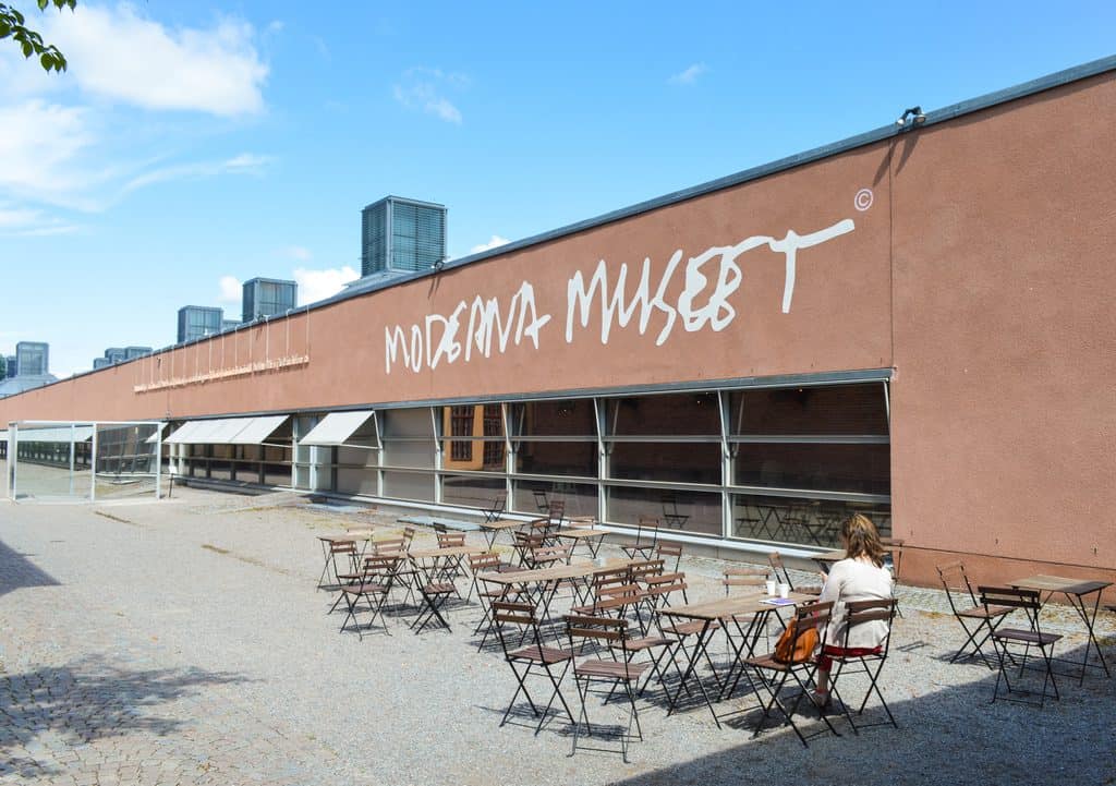 En lång, låg byggnad med rödbrun putsad fasad och texten Moderna Museet i vit handskrift, med takfönster i rad på taket och en uteservering med bord och stolar framför glasfasaden en solig sommardag.