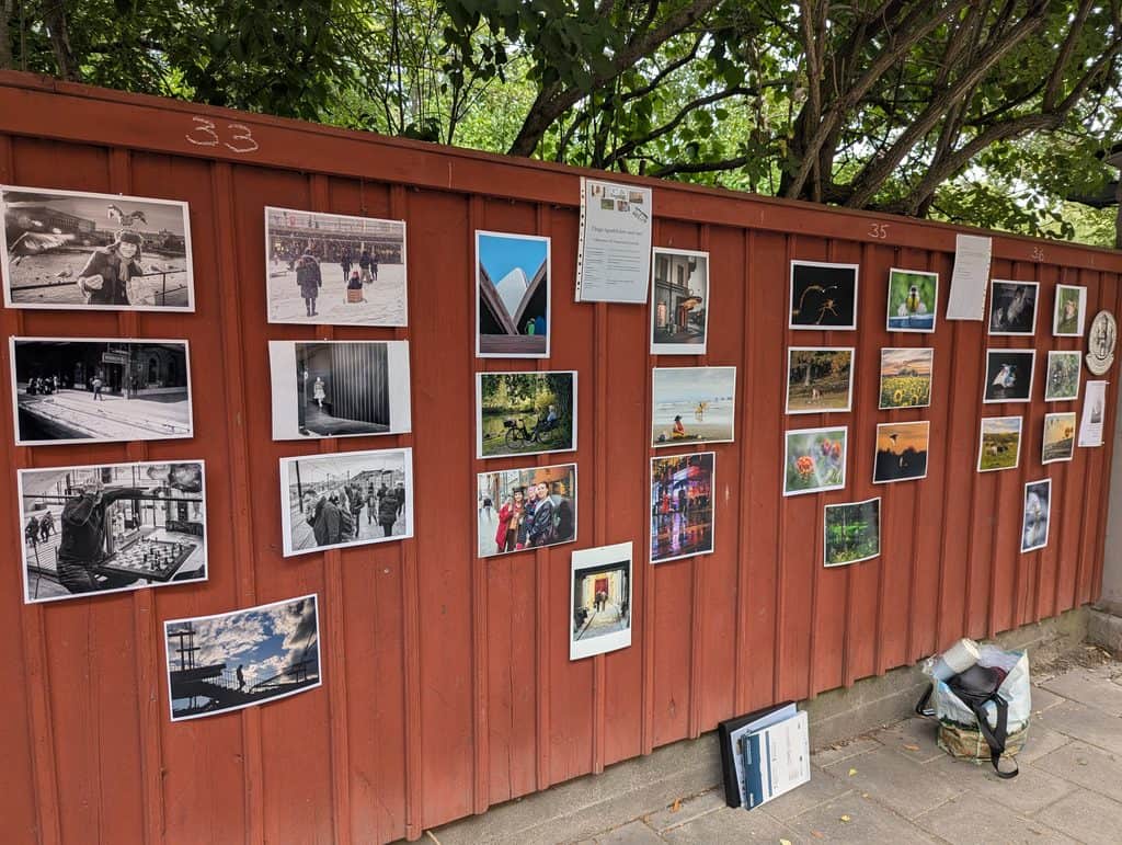 Ett rött träplank i Stockholm med ett fyrtiotal fotografier uppsatta i rader, under lövträd.