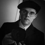 William Molander håller upp sin kamera framför bröstet i ett självporträtt, i basker och runda glasögon.