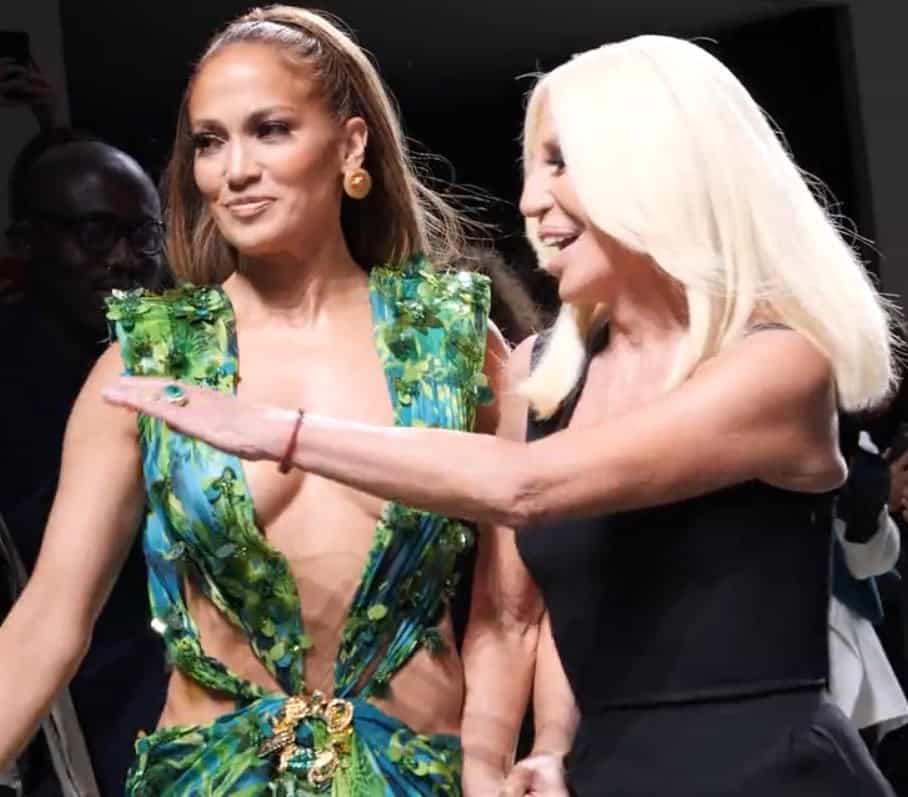 Jennifer Lopez i en ärmlös grön djungelmönstrad klänning med djup urringning och en guldbrosch, bredvid Donatella Versace med långt platinablont hår och svart klänning, som sträcker ut armen mot henne.