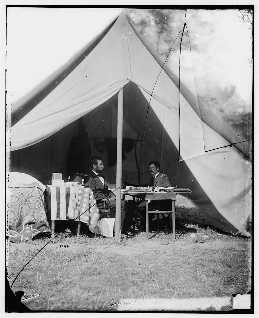Svartvitt fotografi av ett öppet fältält där Abraham Lincoln i mörk kostym sitter vänd mot general McClellan i uniform, som sitter vid ett enkelt skrivbord. Bredvid Lincoln står ett bord draperat med en amerikansk flagga.