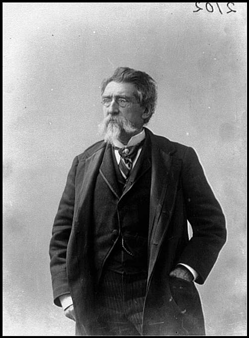 Svartvitt porträtt av Mathew Brady som äldre man i trekvartsfigur, med glasögon, vit mustasch och pipskägg, klädd i mörk rock och väst med händerna i fickorna.