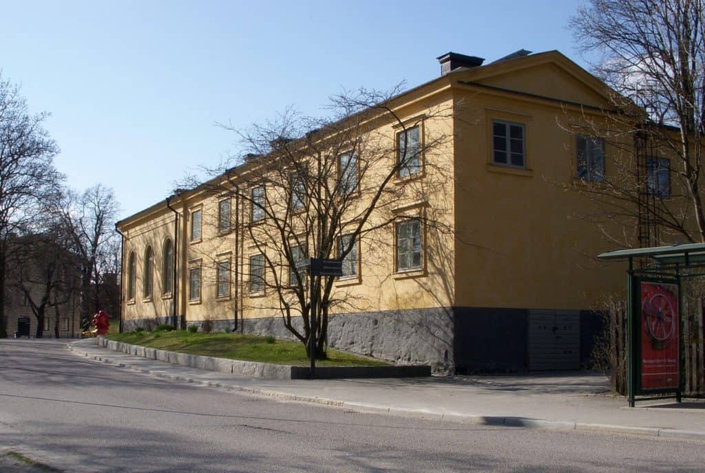 En lång gul byggnad i två våningar med grå stensockel och spröjsade fönster, sedd från gatan en solig vårdag med kala träd framför fasaden och en busskur till höger.