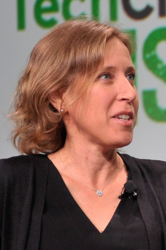 Porträtt av Susan Wojcicki med axellångt ljusbrunt hår, svart tröja och en mygga på kragen, framför en grön och vit skylt från en konferens.