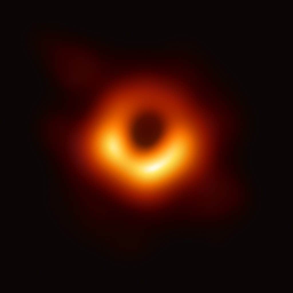 En suddig, glödande ring i orange och gult mot svart bakgrund, med en mörk rund fläck i mitten. Ringen är ljusast på nedre halvan och bleknar uppåt.