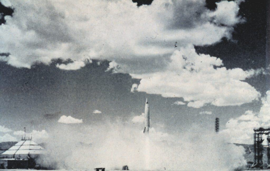 En svartvit bild av en raket som stiger upp från ett platt ökenlandskap med en lång ljus eldsvans under sig och ett moln av damm vid marken. Bergskedjor syns vid horisonten.