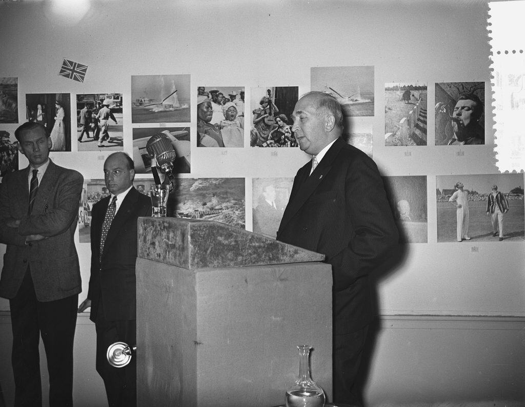 Svartvit bild av en äldre man i mörk kostym som talar vid ett stenklätt podium med mikrofon, medan två yngre män står och lyssnar intill, framför en vägg med uppsatta pressfotografier.